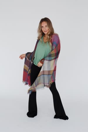A woman stands against a white background wearing a green top, black pants, and the Cozy Plaid Pocket Ruana Wrap - Fuchsia. She looks at the camera with a slight smile, one hand on her hip, standing confidently with legs apart.