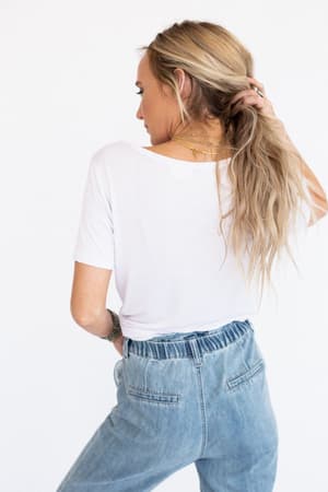 A woman with long blonde hair in a ponytail stands with her back to the camera, wearing the Scoop Neck Bralette Tee in white and high-waisted blue jeans. She holds her hair with one hand and accessorizes with layered necklaces.