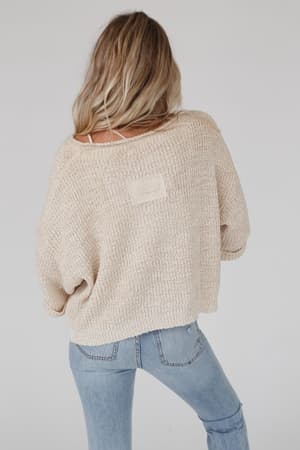 A woman with long, wavy blonde hair faces away, wearing the Just Right Slouchy Sweater in Oatmeal and light blue jeans. The background is plain and light gray.