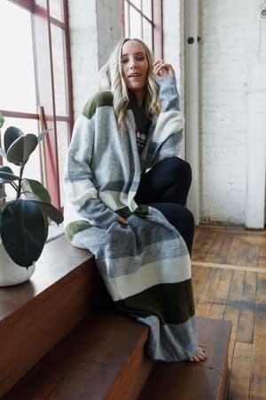 A woman with long blond hair sits barefoot on wooden stairs by large windows, wearing black leggings and The Nest So Cozy Oversized Striped Knit Duster in Olive Multi. A potted plant is beside her.