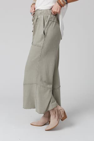 Wearing the Feeling Good Wide Leg Pant in Faded Sage, a white shirt, tan fringe ankle boots, and layered beaded bracelets, a person poses against a light background for an easygoing boho style look.