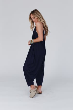 A woman with long blonde hair wears The Perfect Harem Jumpsuit - Navy and beige slip-on shoes, accessorized with colorful bracelets. She stands sideways against a white background, looking down in a relaxed pose.