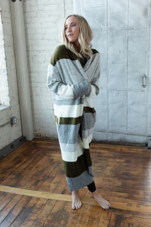 A barefoot blonde woman smiles softly to the side, wearing The Nest So Cozy Oversized Striped Knit Duster in Olive Multi, maxi length, standing by a white brick wall and metal door on wooden floors.