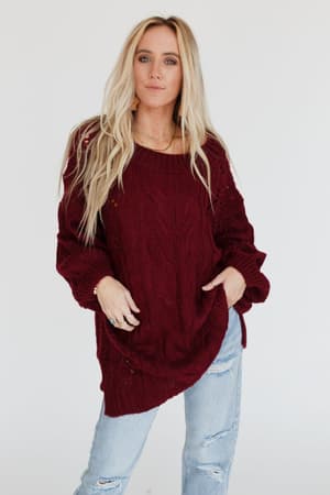 A woman with long blonde hair wears the Golden Hour Bubble Sleeve Sweater Dress in burgundy, standing against a plain white background, looking at the camera with her hands in her pockets.