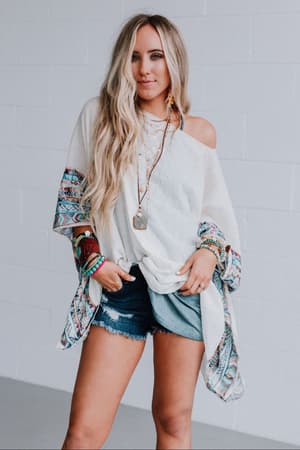 A woman with long blonde hair wears the Fallon Embroidered Sleeve Poncho - Ivory, denim shorts, layered necklaces, and colorful bracelets while standing against a white brick wall.