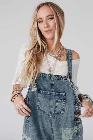 A woman with long blonde hair wears layered necklaces, a white top, colorful beaded bracelets, and the High Hopes Slouchy Denim Overalls - Denim Bandana, standing against a plain light background.