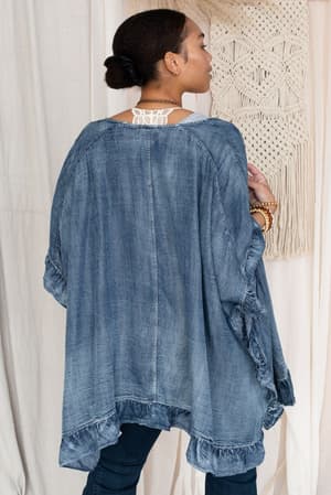 A woman stands with her back to the camera, wearing the Robbie Denim Jacket - Blue, an oversized jacket with ruffle trim. Her hair is tied back, and she wears bracelets. A woven wall hanging appears in the background.