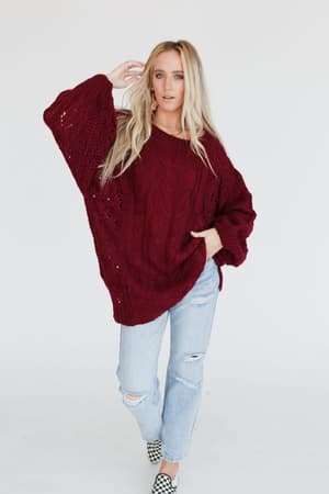 A woman with long blonde hair poses against a white background wearing the Golden Hour Bubble Sleeve Sweater Dress in burgundy. She stands with one hand in her pocket, paired with light ripped jeans and black-and-white checkered slip-ons.