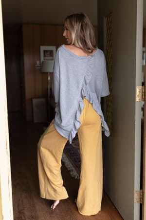 A woman stands barefoot indoors, wearing loose yellow pants and The Nest Must Have High Low Tee in Heather Gray with a ruffled, open back. Facing away from the camera, she's about to step into a warmly lit room.