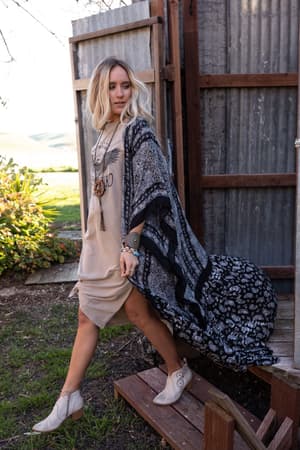 A woman with blonde hair wears a beige dress, layered necklaces, ankle boots, and the Paisley Tapestry Free Flow Duster Kimono in black. She stands outside on wooden steps with greenery and corrugated metal walls behind her.