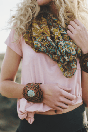 A woman with wavy blonde hair wears a pink knotted shirt, a yellow and blue scarf, and the Aegean Boho Leather Cuff Bracelet - Brown with turquoise stones, plus other decorative bangles. She stands outdoors, face unseen.