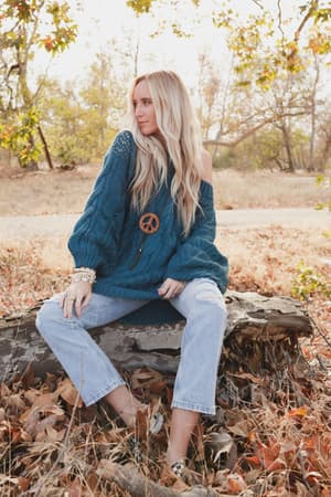 A woman with long blonde hair sits on a fallen log outdoors, wearing the Golden Hour Bubble Sleeve Sweater Dress in teal. Autumn leaves and trees fill the background.