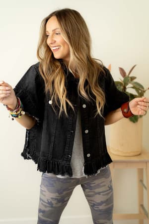 A woman with long wavy hair smiles indoors wearing the Scottie Ruffle Denim Jacket - Black over a gray shirt, camo leggings, and colorful bracelets. A potted plant rests on a small table behind her.