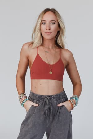 A woman with blonde hair wears the Tattoo Bralette - Rust, paired with gray drawstring pants, layered necklaces, and beaded bracelets. She stands against a plain light background with her hands in her pockets.
