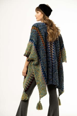 A woman with long, wavy hair wears a black headband, flared gray pants, and the Desert Dreams Crochet Ruana – Cobalt, featuring vibrant zigzag patterns and tasseled corners that enhance her boho style against a plain light background.
