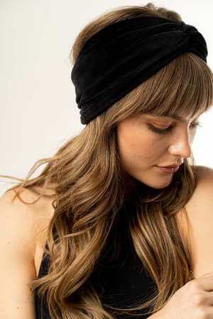 A woman with long, wavy light brown hair wears the Everyday Velvet Headband in Black, featuring subtle criss-cross detailing, paired with a sleeveless black top as she looks down against a neutral background.