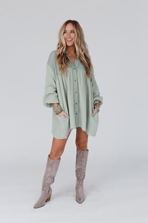 A woman with long blonde hair wears the Sunday Kind Of Love Dress in sage, an oversized light green button-up shirt dress with pockets, styled with taupe knee-high boots and colorful bracelets, smiling against a plain white background.