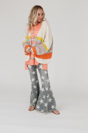 A woman stands against a plain background wearing the Retro Babe Striped Cardigan - Cream over a peach graphic hoodie, distressed star-print flared jeans, and sandals. She looks slightly to the side with a relaxed posture.