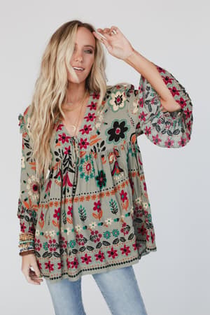A woman with long blonde hair smiles, wearing the Leona Floral Print Tunic in Sage with billowy sleeves and light blue jeans, posing against a plain white background.