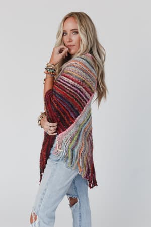 A woman with long blonde hair poses sideways against a plain light background, wearing the Madeleine Poncho - Multi and distressed light blue jeans, with one hand near her face.