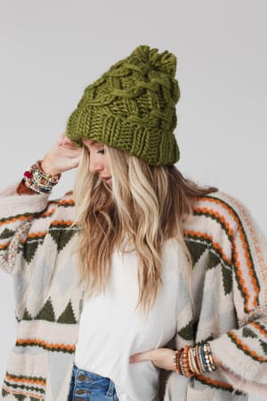 A woman wearing a green oversized chunky knit beanie with a pom-pom design, paired with a colorful patterned shawl and a white top.