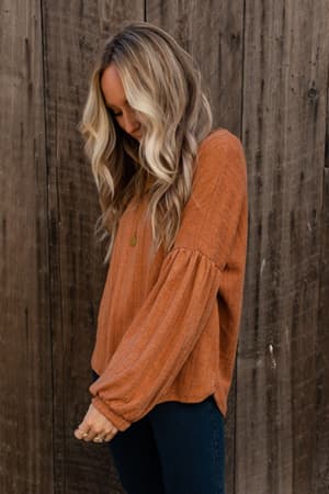A woman with long wavy blonde hair stands in front of a wooden wall, looking down. She is wearing The Nest Sweet Heart Balloon Sleeve Knit Sweater in burnt orange and dark jeans.