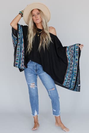 A woman with long blonde hair poses barefoot against a plain background, wearing ripped blue jeans and the Fallon Embroidered Sleeve Poncho in black, styled with a wide-brimmed hat, layered necklaces, and bracelets.