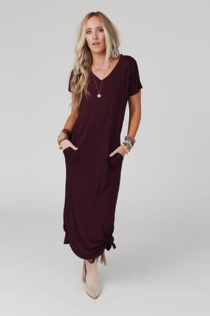A woman stands against a plain background wearing the Call It Comfort Tee Dress - Plum, a short-sleeved, relaxed-fit midi dress with side pockets. She accessorizes with boho jewelry and beige ankle boots, her long blonde hair loose.