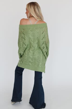 A woman with long blonde hair stands facing away, wearing the Golden Hour Bubble Sleeve Sweater Dress in light green over a gray top and dark blue flared jeans, posed against a plain light background.