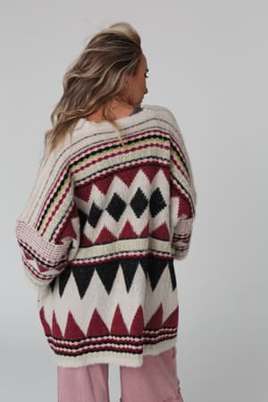 A person with wavy blonde hair faces away, wearing the Keep It Up Oversized Cardigan - Berry, a cozy chunky knit with geometric black, red, green, and white patterns, paired with pale pink pants against a plain light background.