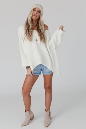 A woman wears the Dana Oversized Diamond Texture Sweater in ivory, styled with denim shorts, beige ankle boots, and a white knit beanie, standing against a plain light background.