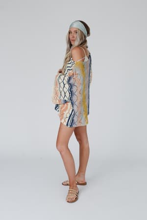 A woman with long, light hair wears the Rainbow Shores Knit Sweater - Multi, paired with a blue headband and pearl-detailed sandals. She stands sideways, looking at the camera against a plain white background.