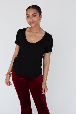 A woman stands against a white background, wearing the Scoop Neck Bralette Tee - Black with red velvet pants and gold jewelry. Her hair is pulled back and she smiles slightly—a chic example of boho tops for women.