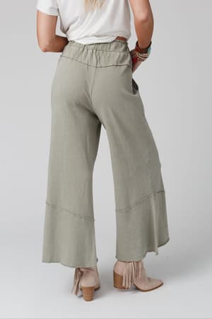 Wearing the Feeling Good Wide Leg Pant in Faded Sage, a white top, beige fringe ankle boots, and colorful bracelets, this person radiates relaxed boho style from the back against a plain light background.