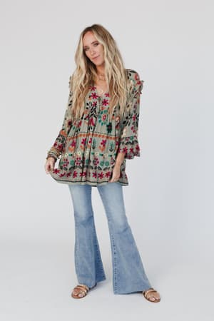 A woman with long blonde hair wears the Leona Floral Print Tunic in Sage, paired with light blue flared jeans and sandals, standing against a white background and smiling gently while holding her blouse.