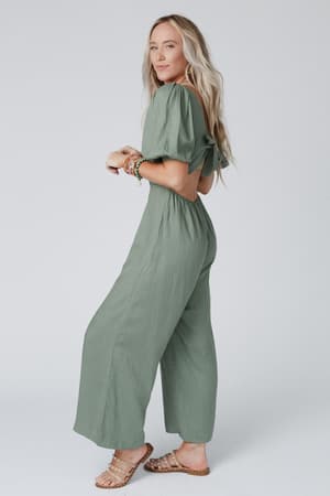 A woman with long blonde hair wears the Weekender Puff Sleeve Jumpsuit in olive, paired with tan studded sandals and beaded bracelets, posing sideways against a plain light background.