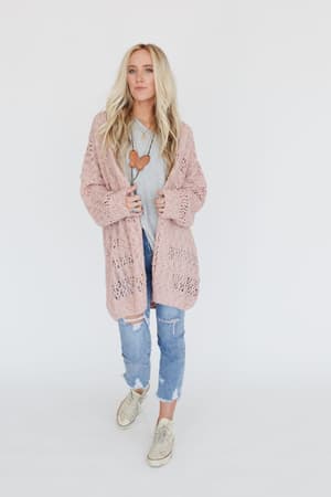 A woman with long blonde hair wears the Cara Oversized Cardigan in pink over a gray shirt, paired with ripped blue jeans and white sneakers, standing against a white background.