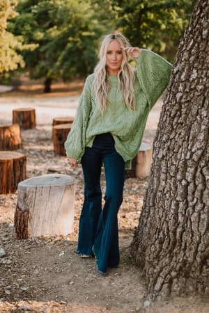 A woman with long blonde hair stands outdoors by a tree, wearing the Golden Hour Bubble Sleeve Sweater Dress in light green. Tree stumps surround her, and lush green trees form the background.