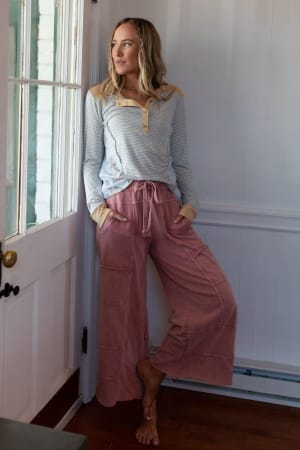 A woman stands indoors by a white door, barefoot and leaning against the wall, wearing loose Lucky One Wide Leg Pant in mauve with a long-sleeve striped shirt featuring beige accents, looking outside through the glass.