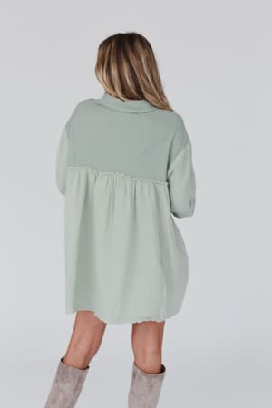 A woman with long blonde hair stands with her back to the camera, wearing the Sunday Kind Of Love Dress - Sage and light khaki knee-high boots, set against a plain, light background.