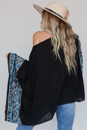 A woman with long blonde hair in a beige hat and blue jeans faces away, wearing the Fallon Embroidered Sleeve Poncho in black, featuring colorful patterned sleeves.