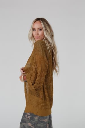 A woman with long blonde hair wears the Brenden Knit Netted Cardigan in brown and camouflage pants, standing sideways and looking at the camera with a neutral expression against a plain light background.