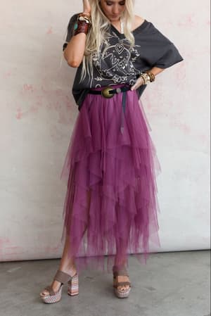 A woman in a loose graphic tee, Boho Ballerina Skirt - Mauve in layered tulle, platform sandals, and bracelets stands against a light pink and white textured wall, touching her hair with one hand.