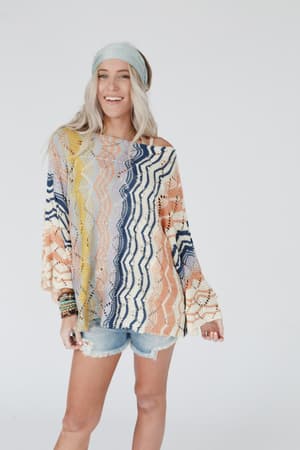 A woman with long blonde hair wearing a blue headband, denim shorts, beaded bracelets, and the Rainbow Shores Knit Sweater - Multi stands smiling against a plain white background.