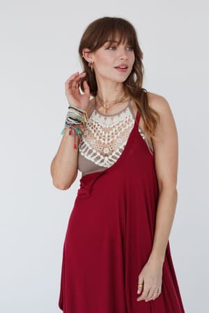 A woman with long brown hair wears a sleeveless red dress over the Crochet High Neck Bralette - Mocha, accessorized with layered necklaces, hoop earrings, and colorful beaded bracelets. She poses against a plain white background.