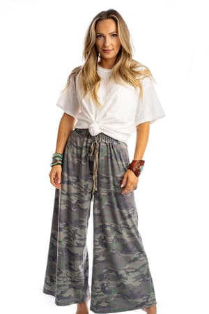 A woman with long blonde hair and bracelets wears the Calling All Camo Lounge Pants - Olive, featuring a loose wide-leg silhouette. She pairs them with a white knotted t-shirt and stands facing forward against a white background.