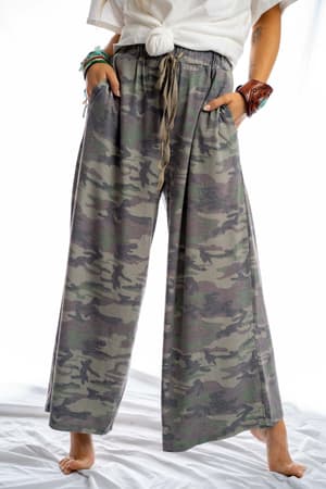 Barefoot on a white surface, a person wears the Calling All Camo Lounge Pants - Olive, featuring a loose fit, wide leg silhouette, and pockets. A tied white shirt and bracelets complete the look; head and upper body are mostly out of frame.
