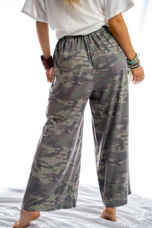 A person stands barefoot on a white surface, back to the camera, wearing loose Calling All Camo Lounge Pants - Olive and a white t-shirt, with colorful bracelets visible on one wrist.