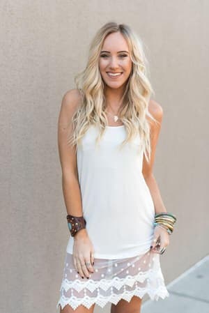A smiling woman with long, wavy blonde hair wears the Layering Love Lace Slip - Ivory, a sleeveless white dress with sheer lace details and subtle polka dots. She adds multiple bracelets and a brown cuff, standing against a plain beige wall.