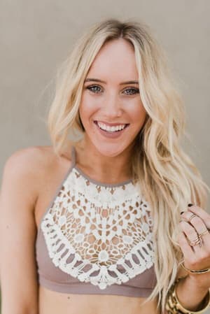 A smiling woman with long, wavy blonde hair wears the Crochet High Neck Bralette in Mocha and multiple rings, standing against a plain, light-colored background.